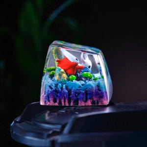 May include: A custom artisan keycap featuring a miniature diorama. The keycap showcases two figures in a colorful, layered scene with blue, purple, and green elements. The figures are a red character and a white character. The keycap is encased in clear resin.