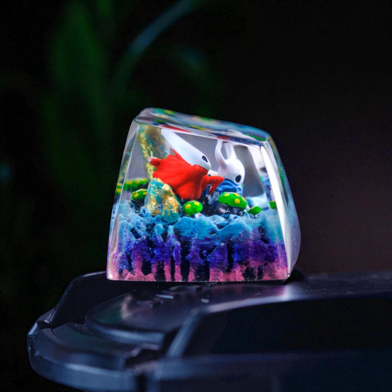 May include: A custom artisan keycap featuring a miniature diorama. The keycap showcases two figures in a colorful, layered scene with blue, purple, and green elements. The figures are a red character and a white character. The keycap is encased in clear resin.