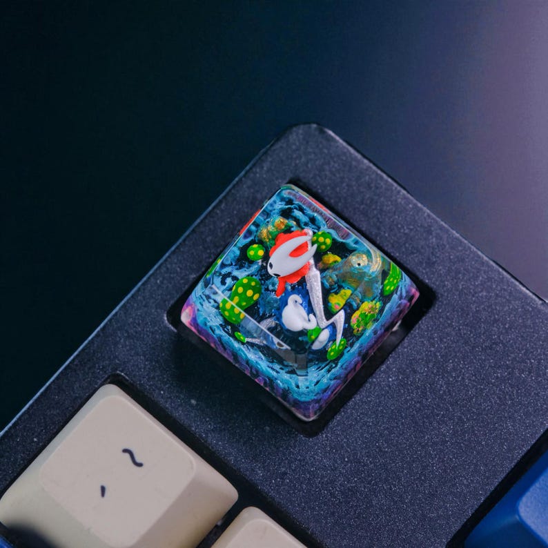 May include: A custom artisan keycap featuring a colorful underwater scene. The keycap has a clear resin base with a design of a red and white fish, green plants, and other aquatic elements. The keycap is square and sits on a black keyboard.