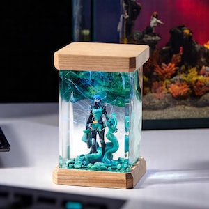 May include: A rectangular resin art piece featuring a figurine inside a clear, glass-like enclosure. The figurine is a character in a teal and black outfit. The base and top are made of light-colored wood. The resin has teal and white swirls.