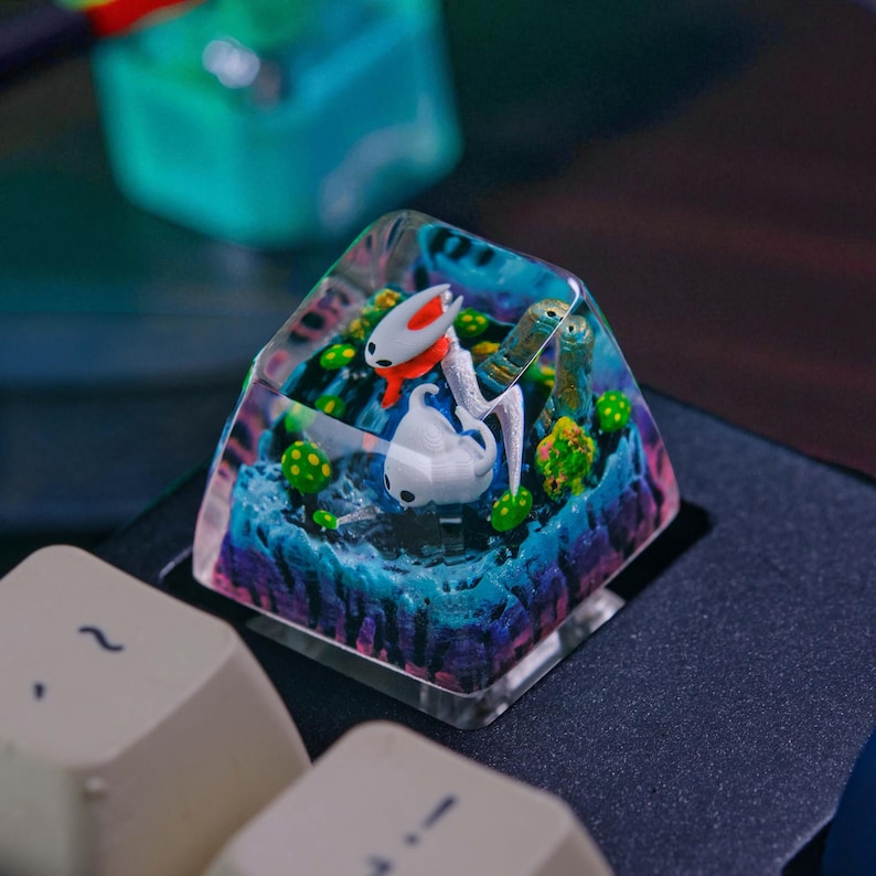 May include: A custom artisan keycap featuring a miniature landscape with a white and red character and a white character. The keycap is clear, with a blue, green, and purple landscape design. The keycap is square and sits on a black keyboard.