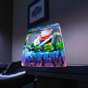 May include: A clear, rectangular resin art piece featuring a miniature diorama. The diorama includes a white and red figure, blue and green elements, and a colorful, textured base with shades of purple, blue, and green.