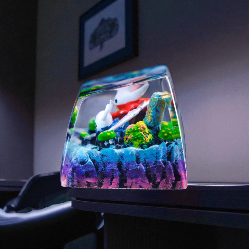 May include: A clear, rectangular resin art piece featuring a miniature diorama. The diorama includes a white and red figure, blue and green elements, and a colorful, textured base with shades of purple, blue, and green.