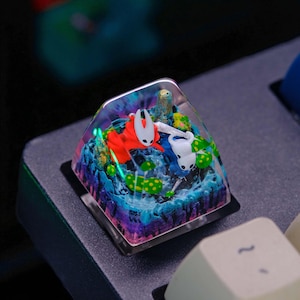 May include: A custom keycap featuring a miniature diorama with two cartoon characters in a colorful landscape. The keycap has a clear, transparent top, and the scene includes blue, green, and purple elements. The keycap is square and sits on a black keyboard.