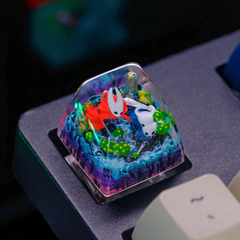 May include: A custom keycap featuring a miniature diorama with two cartoon characters in a colorful landscape. The keycap has a clear, transparent top, and the scene includes blue, green, and purple elements. The keycap is square and sits on a black keyboard.