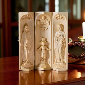 Op de afbeelding: Een houten drieluik met gebeeldhouwde religieuze figuren. De panelen tonen Ave Maria, de Heilige Drie-eenheid en St. Joseph. De gravures zijn gedetailleerd en geplaatst in gewelfde frames. Het drieluik is gemaakt van lichtgekleurd hout.