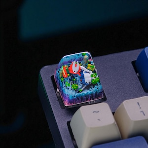 May include: A custom keyboard keycap featuring a colorful, detailed scene with two characters, one red and one white, in a watery environment. The keycap has a clear, glossy finish and is set on a dark gray keyboard.