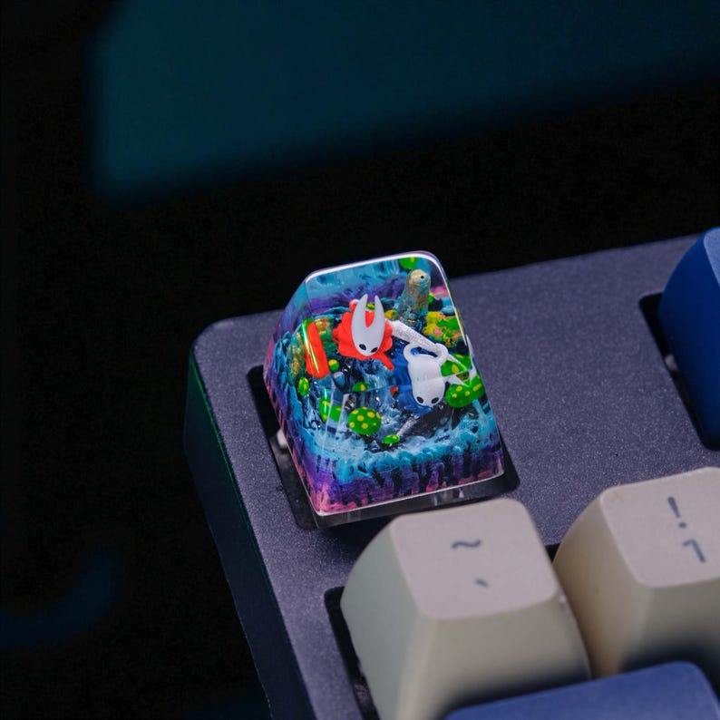 May include: A custom keyboard keycap featuring a colorful, detailed scene with two characters, one red and one white, in a watery environment. The keycap has a clear, glossy finish and is set on a dark gray keyboard.