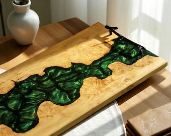 May include: Wooden serving tray with a river of green epoxy resin. The tray is rectangular with a light wood grain and dark metal handles. A white vase is in the background.