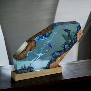 May include: A fantasy resin sculpture with a castle, dragons, and a tree encased in a clear block. The piece has a wooden base and accents, with blue and purple tones. The design creates a magical, whimsical aesthetic, suitable for display.