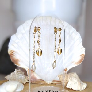 Peut inclure: Boucles d'oreilles pendantes dorées avec une petite perle, un charme coquillage doré et une chaîne dorée. Les boucles d'oreilles sont présentées sur un support en acrylique transparent avec un fond en coquillage.