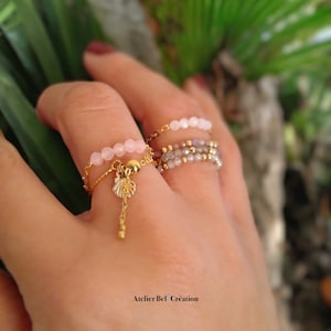 May include: Several delicate rings are displayed on a hand. One ring has small pink beads and a gold charm with a leaf design. Other rings feature small beads in shades of pink and gold. The rings are on a gold chain. The text "Atelier Bel Création" is at the bottom.