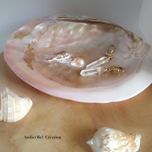 Peut inclure: Une paire de boucles d'oreilles en or avec des pendentifs en cristal de quartz clair, reposant sur une coquille de nacre rose et blanche. Les boucles d'oreilles sont présentées sur une surface en bois avec deux coquillages.