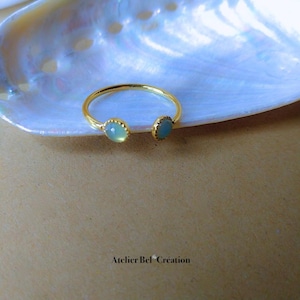 Peut inclure: Une bague ouverte dorée avec deux pierres précieuses rondes bleu clair. La bague est posée sur un fond nacré et une surface marron. Le texte "Atelier Bel Création" est visible.