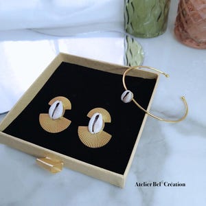 May include: A jewelry set featuring gold-toned earrings and a bracelet, each adorned with a white seashell. The earrings have a fan-like design. The set is presented in a beige box with a black interior.
