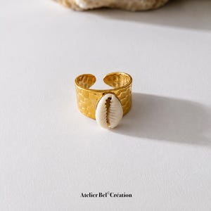 Puede incluir: Un anillo de tono dorado con una superficie texturizada y martillada, con un diseño de banda abierta. Centrado en la parte frontal hay un caracol cauri blanco de forma ovalada. El texto Atelier Bel Création aparece impreso en negro.