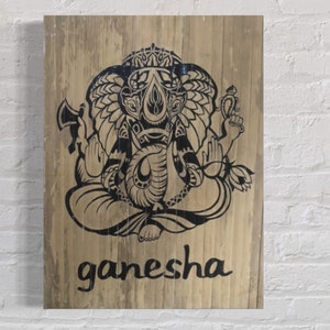 May include: A wooden sign featuring a black and white illustration of Ganesha, the Hindu god of beginnings, prosperity, and good fortune. The sign has the word "ganesha" written below the illustration.