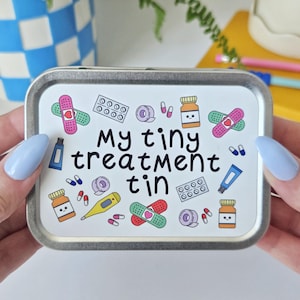 May include: A rectangular metal tin with a white lid displaying the text "My tiny treatment tin". The lid is decorated with colourful illustrations of plasters, pills, medicine bottles, and a thermometer. The tin is held by hands with light blue nails.