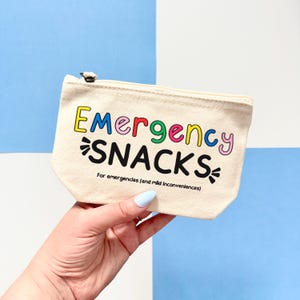 May include: A beige canvas zippered pouch featuring the multicoloured text Emergency in a rounded font, the black text SNACKS in a bold font, and the smaller black text For emergencies (and mild inconveniences) printed on the front.