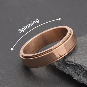 Può includere: Un anello color oro rosa con una fascia esterna rotante. L'anello è posto su uno sfondo grigio scuro. La parola "Spinning" è scritta sopra l'anello con una freccia che indica la direzione della rotazione.
