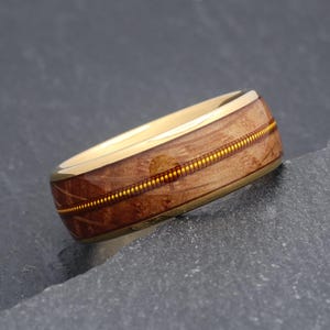 May include: A gold ring featuring a wooden inlay and a thin, dotted gold band. The wood has a warm, brown hue with a subtle grain pattern. The ring is set against a dark grey background.