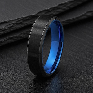 May include: A ring featuring a brushed black exterior finish and a polished, vibrant blue interior lining. The band has a flat profile with beveled edges, showcasing a distinct contrast between the dark outer surface and the bright blue inner band.
