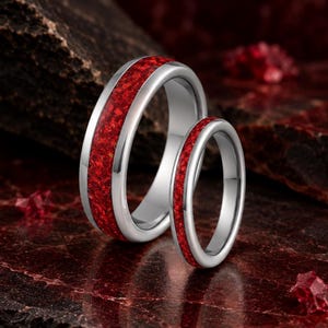 May include: Two silver-toned metal rings featuring a central inlay of crushed red material. The wider ring stands upright behind a thinner ring, both displaying a polished metallic finish and a vibrant, textured red band running along the centre.