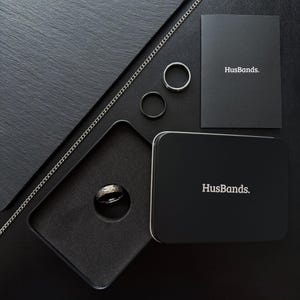 May include: A black ring in a black velvet box with a black chain and two silver rings. The box and the chain have the word "HusBands." printed on them.
