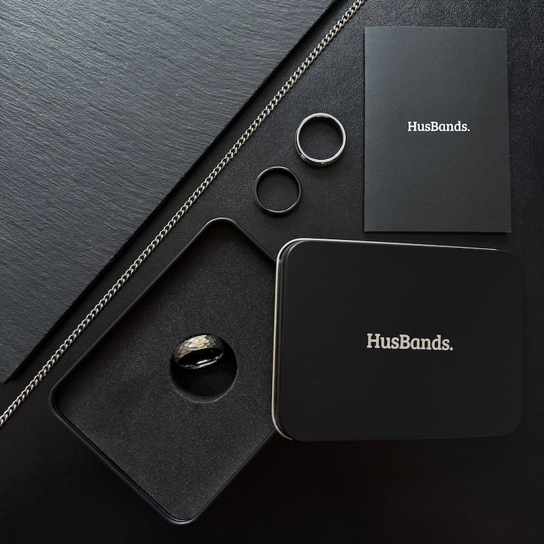 May include: A black ring in a black velvet box with a black chain and two silver rings. The box and the chain have the word "HusBands." printed on them.