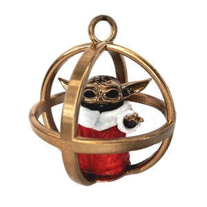 Puede incluir: Un adorno dorado en forma de esfera con una pequeña figura de un personaje de la franquicia Star Wars en su interior. El personaje lleva un atuendo rojo y blanco y está de pie en una estructura en forma de jaula.