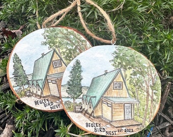 May include: Two round wooden ornaments featuring hand-painted illustrations of an A-frame cabin with a green roof and trees. The text "BEGLEY BIRD NEST" is written on both, with one including "EST 2022". Each ornament hangs from a brown twine loop.