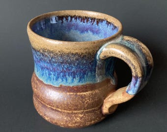 May include: A handmade ceramic mug with a brown, blue, and purple glaze. The mug has a unique, slightly rounded shape and a textured handle.