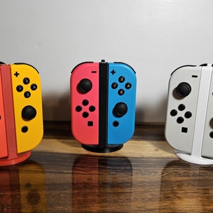 May include: Three Nintendo Switch Joy-Con controllers in different colors: orange and yellow, pink and blue, and white. The controllers are attached to a stand.