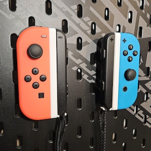 May include: Two Nintendo Switch Joy-Con controllers, one red and white and one blue and white, are hanging on a pegboard wall. Both controllers have a black strap attached.