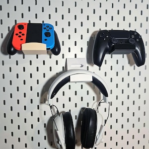 May include: A white pegboard with a white headset, a black and white video game controller, and a red, blue, and white video game controller hanging on it.