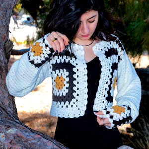 Puede incluir: Un cárdigan de crochet blanco y negro con detalles marrones. El cárdigan tiene un patrón cuadrado y es corto.