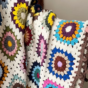 May include: A crocheted throw blanket with a colorful floral pattern. The blanket is made of white yarn with multi-colored squares featuring different floral designs.