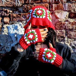 May include: Red, white, and green crochet fingerless gloves with a granny square pattern. The gloves are worn by a person with black nail polish.