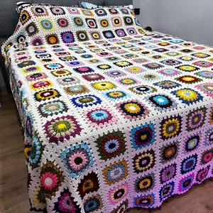 May include: A handmade, colorful crochet blanket with a patchwork design. The blanket features a white background with numerous squares in various colors, including pink, blue, yellow, and green. The blanket is draped over a bed, showcasing its intricate pattern.