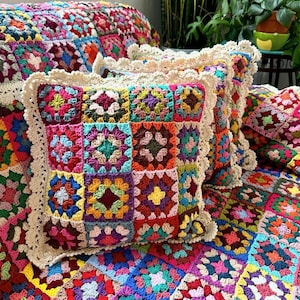 May include: A square throw cushion featuring a colourful granny square crochet pattern in various shades of red, blue, yellow, green, and purple. The cushion is edged with a cream-coloured scalloped crochet border and rests on a matching blanket.