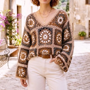 Könnte beinhalten: Ein langärmeliger Häkelpullover mit V-Ausschnitt und einem sich wiederholenden Quadratmuster in Creme-, Braun- und Dunkelgrautönen. Der Pullover wird mit einer weißen Hose, einem braunen Ledergürtel mit silberner Schnalle und einer Goldkette kombiniert.