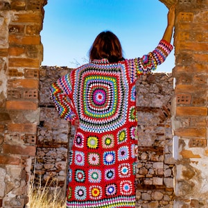 May include: A long, colorful crochet cardigan with a geometric pattern. The cardigan is made of a variety of colors, including red, pink, blue, green, yellow, and black. The cardigan is worn by a person standing in a doorway.