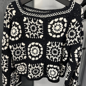 May include: Black and white crochet sweater with a geometric pattern. The sweater has a round neckline and long sleeves.
