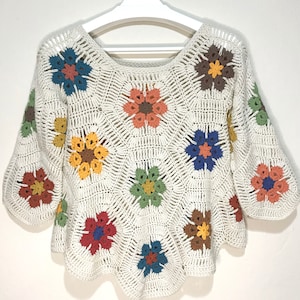 Puede incluir: Un suéter de crochet blanco con un patrón floral en tonos de azul, verde, amarillo, naranja y marrón.