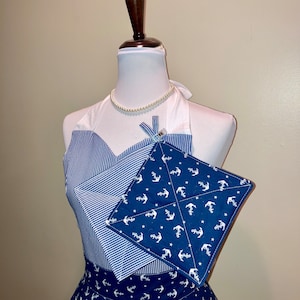 Puede incluir: Un delantal a rayas azules y blancas con un agarrador de olla azul con anclas y estrellas blancas. El delantal tiene un top blanco de halter y un collar de perlas blancas.