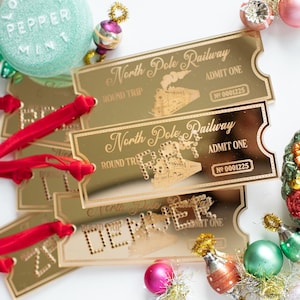 May include: Gold-toned acrylic North Pole Railway tickets with red velvet ribbons. Each ticket features a steam train illustration, the text North Pole Railway, Admit One, Round Trip, and number 0001225, surrounded by small colorful Christmas ornaments.