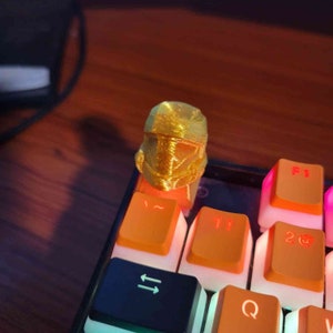May include: A gold-colored, 3D-printed helmet keycap sits atop an orange keyboard key. The keycap is detailed with lines and ridges, resembling a futuristic helmet. The keyboard keys are orange and black.