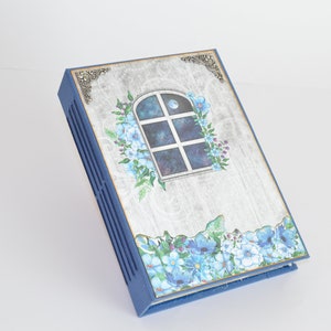 Op de afbeelding: Een blauw journal met een witte houten omslag met een raam met een maan en sterren. Het raam is omringd door blauwe bloemen en groene bladeren. Het journal heeft een decoratief metalen hoekstuk.