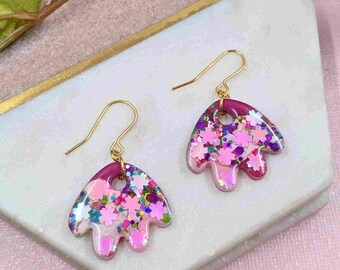 Könnte beinhalten: Ein Paar Ohrhänger mit floralem Design. Die Ohrringe haben einen goldfarbenen Haken und einen klaren, blütenblattförmigen Anhänger, der mit rosa und lila Glitzer und blütenförmigen Konfetti gefüllt ist. Die Oberseite des Anhängers ist tiefmagenta.