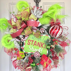 May include: A Christmas wreath with a green, red, and white color scheme. It features a sign that says "stunk! STANK! stunk!" and is decorated with a red and white striped bow, candy canes, and a green and red ornament.